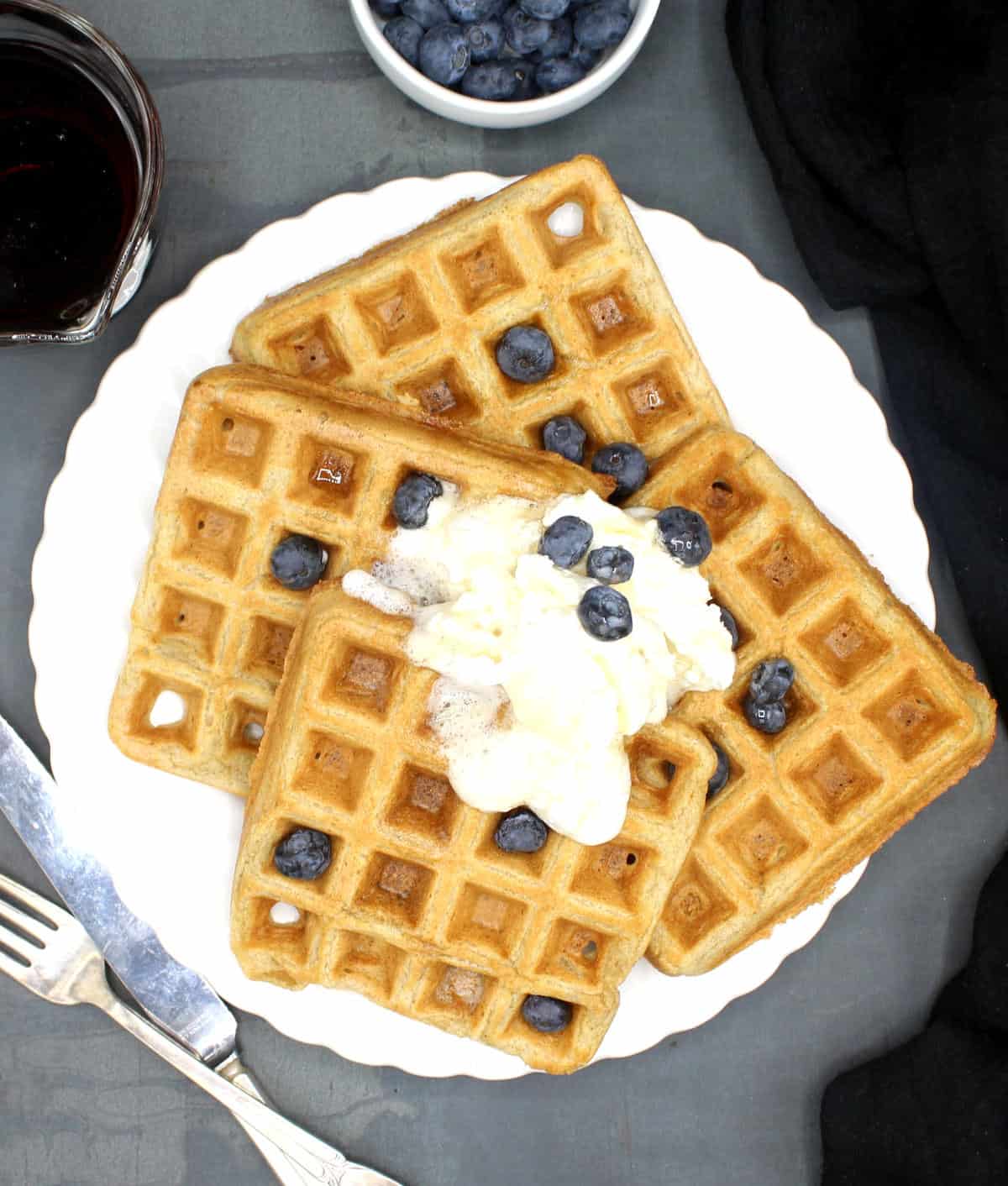 A white plate with four vegan waffles topped with blueberries and vegan whipped cream with maple syrup and blueberries on the side.