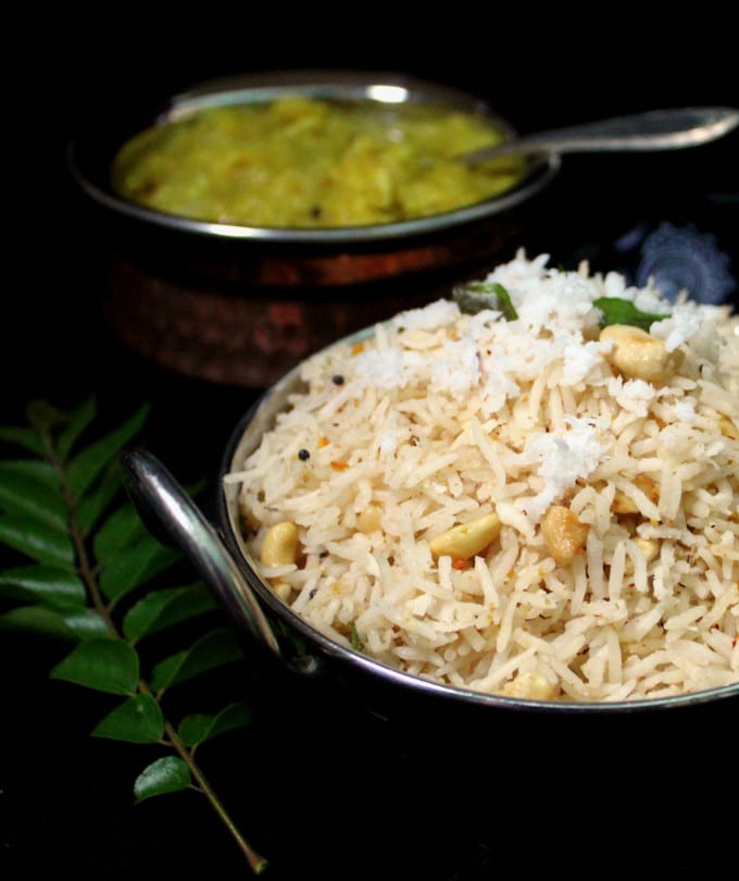 Photo of Coconut rice in a steel bowl with a vegetable side in background and curry leaves next to it.