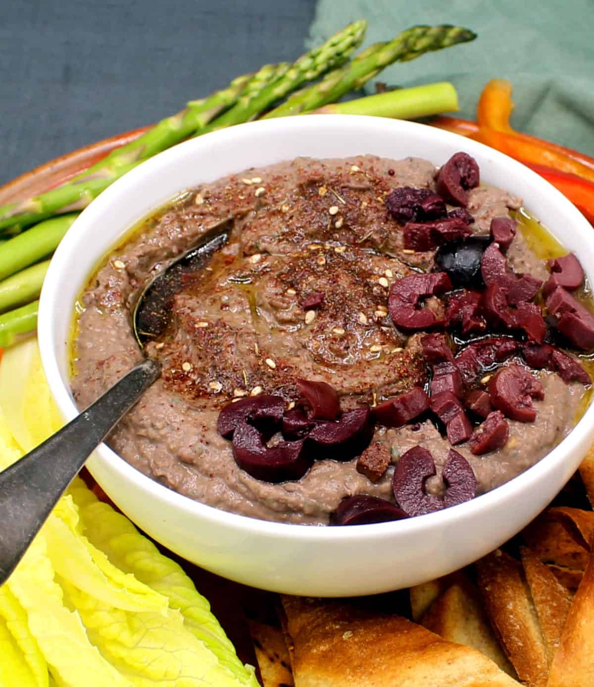 Black bean hummus in white bowl with spoon surrounded by vegetable crudites and pita wedges.