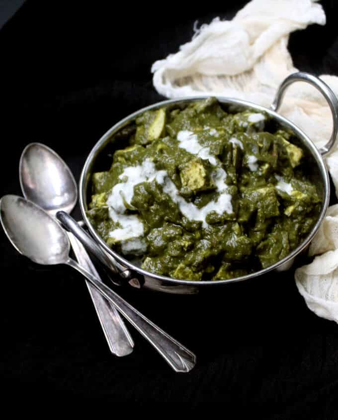 Photo of Palak Paneer with tofu or Saag Paneer in a steel karahi or serving bowl with two spoons on the side against a black background