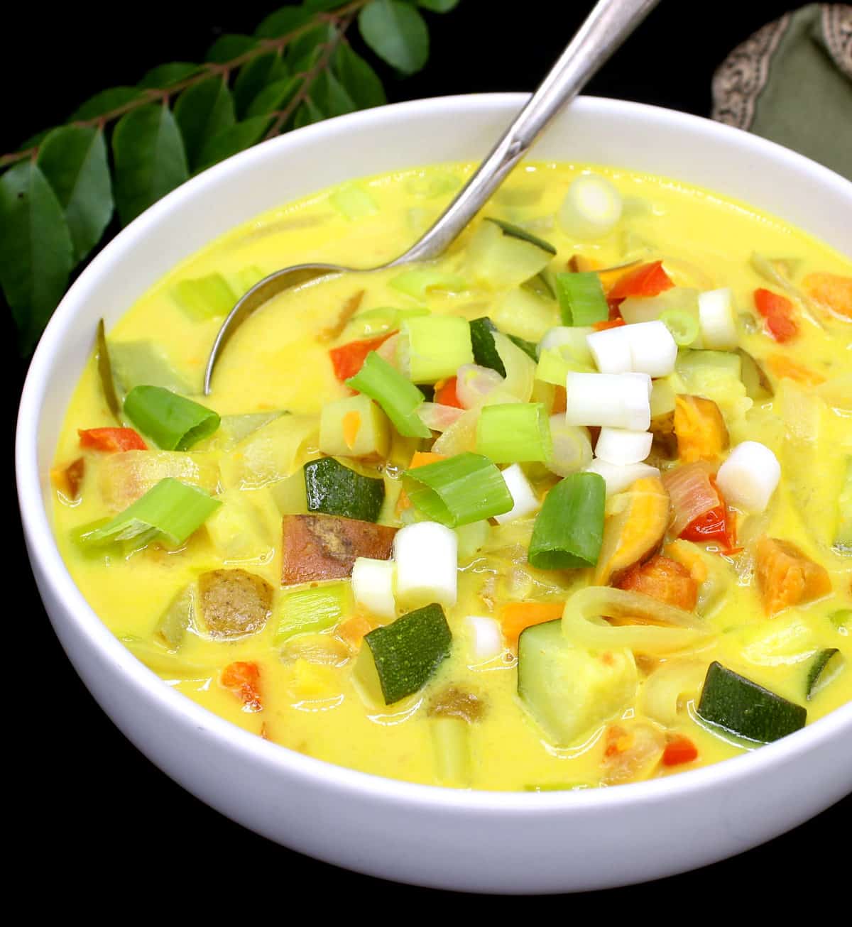 Close up of a white bowl with colorful vegetable moilee with scallions, bell peppers, tomatoes, potatoes, sweet potatoes and zucchini with a spoon and a green napkin and curry leaves