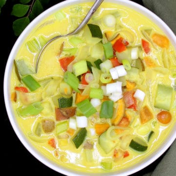 A white bowl with colorful vegetable moilee with scallions, bell peppers, tomatoes, potatoes, sweet potatoes and zucchini with a spoon and a green napkin and curry leaves