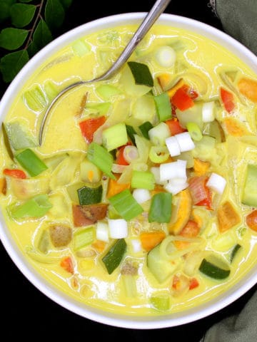A white bowl with colorful vegetable moilee with scallions, bell peppers, tomatoes, potatoes, sweet potatoes and zucchini with a spoon and a green napkin and curry leaves