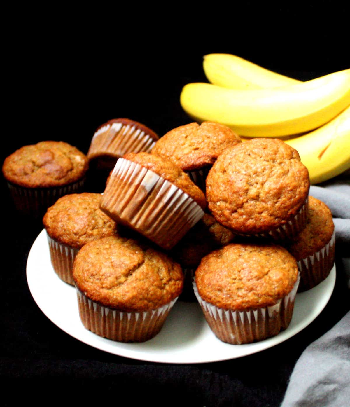 Vegan Banana Muffins stacked on a white plate with ripe bananas in the background.