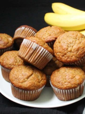 Vegan Banana Muffins stacked on a white plate with bananas in the background