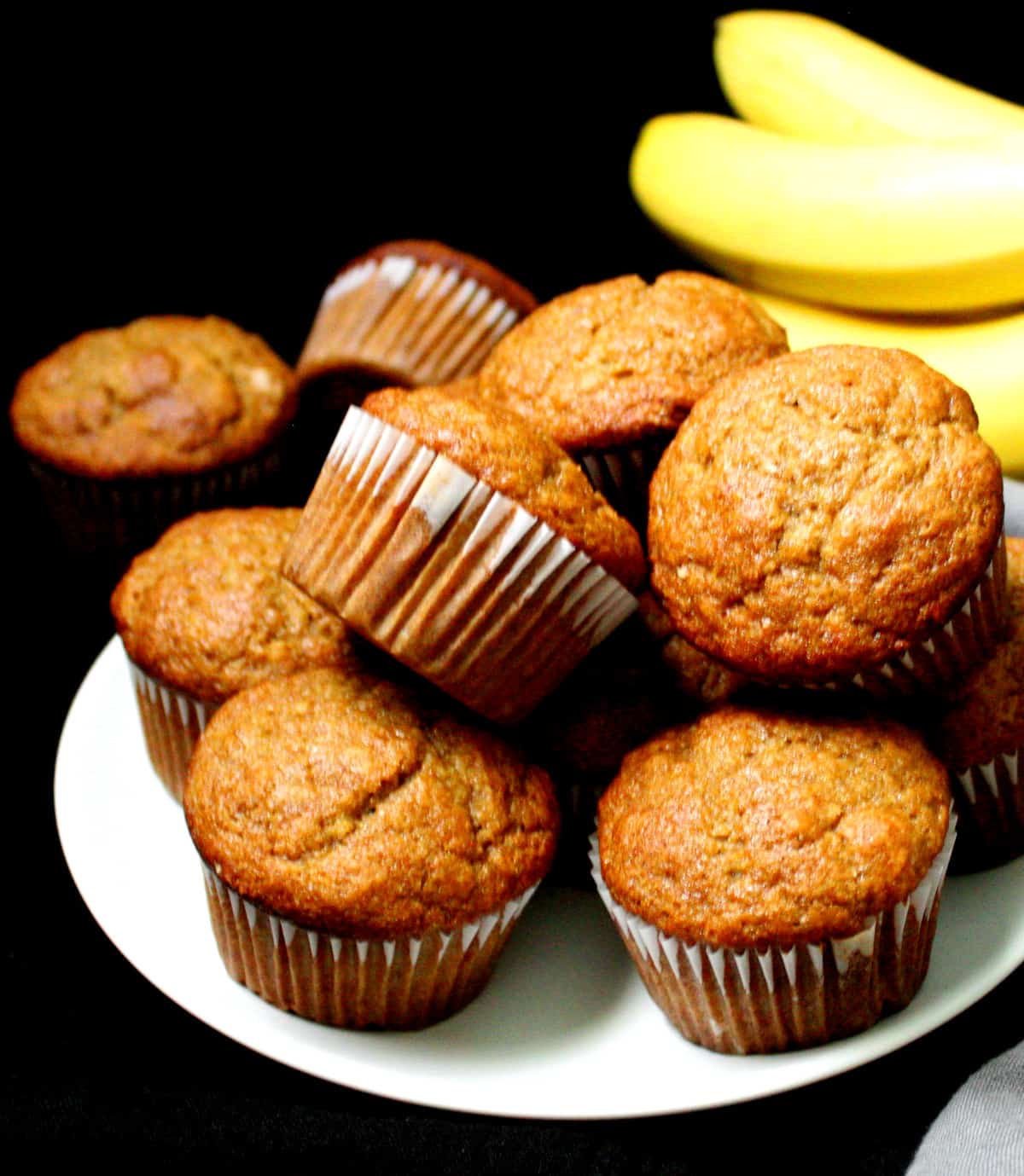 A stack of vegan banana muffins on a white plate with a bunch of yellow bananas in the background.