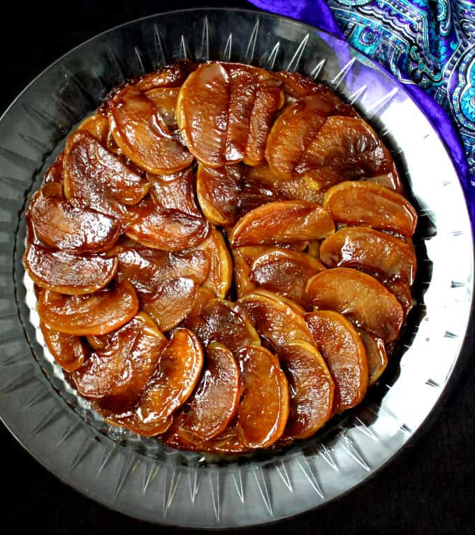 Top short of a glossy vegan Tarte Tatin on a glass crystal plate with blue silk fabric in the background.