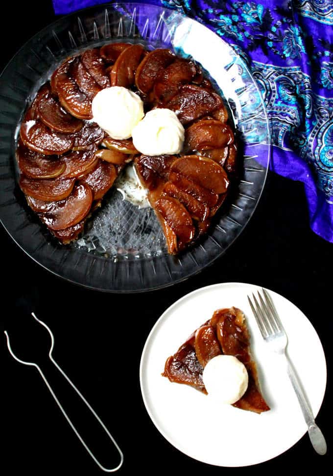 Top shot of a vegan tarte tatin with scoops of ice cream on a glass platter and a white plate with a slice of the apple tart next to it