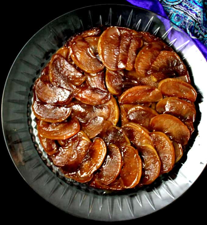 Top shot of a vegan tarte tatin with the amber apple slices arranged in a circular pattern, on a glass platter against a black background