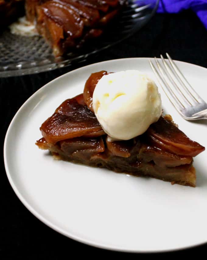A close up shot of a slice of vegan tarte Tatin on a white plate with a scoop of vegan vanilla ice cream on top and a fork on the side