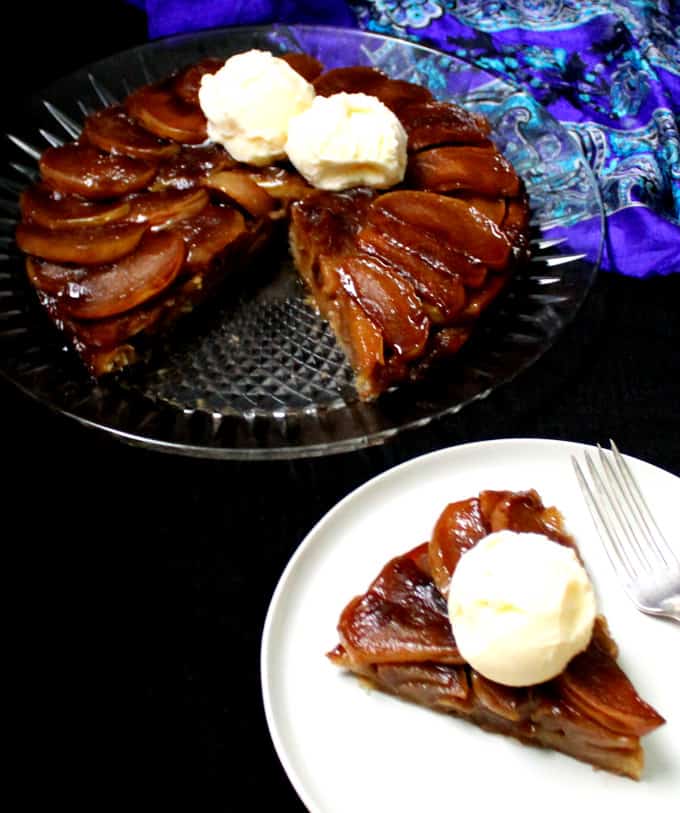 Front shot of a slice of vegan Tarte Tatin with the whole tart in the background on a glass plate with scoops of vegan vanilla ice cream on top