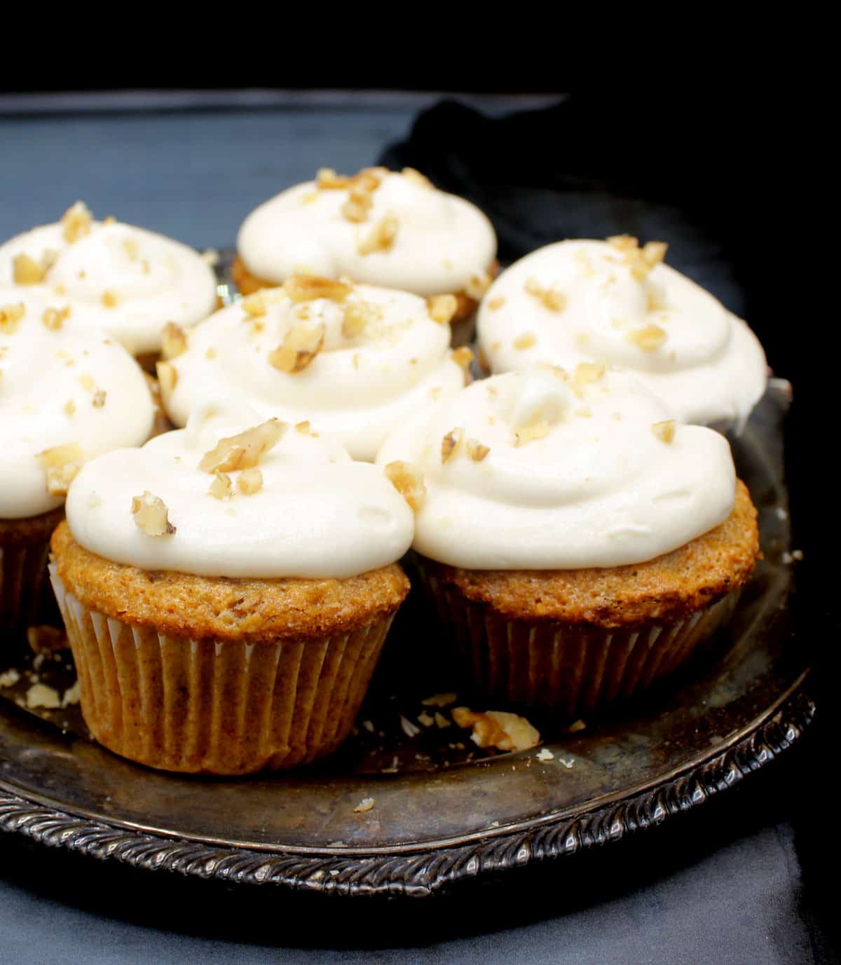 Close front shot of vegan carrot cupcakes with cream cheese frosting and walnuts scattered on top in a silver plate.