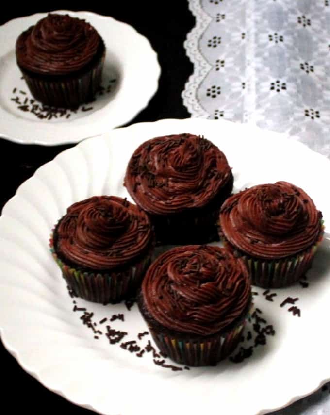 Four vegan chocolate cupcakes with buttercream frosting on a white plate with a single cupcake in the background and a lace napkin.