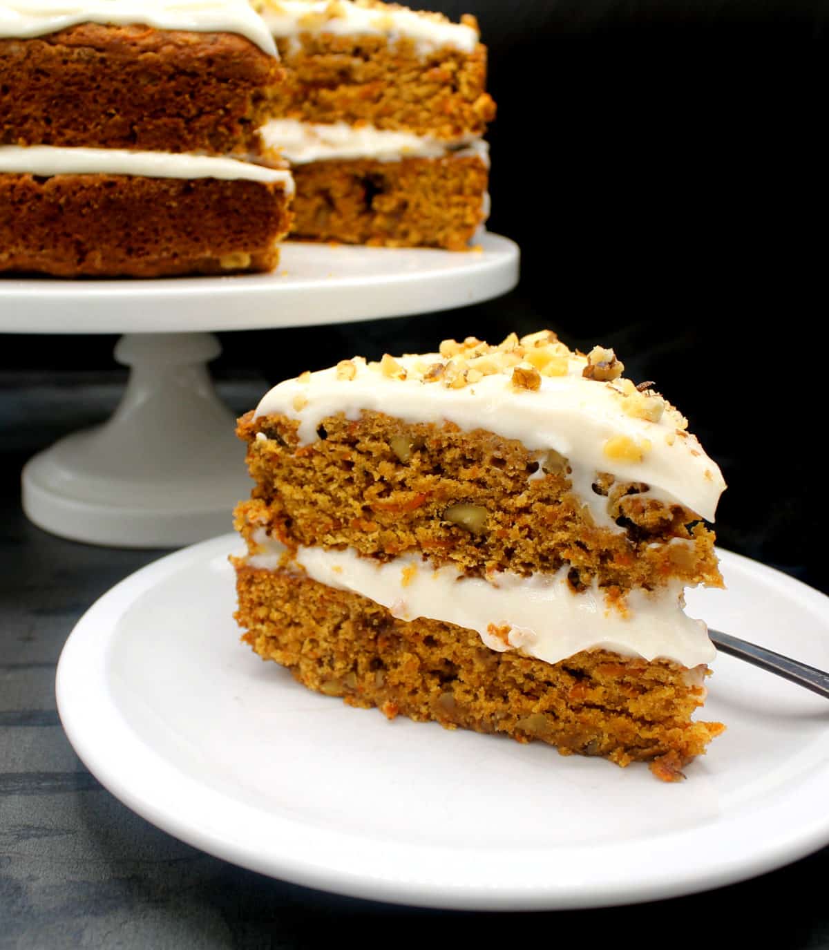 Slice of vegan carrot cake on white plate with cake on cake stand in background.