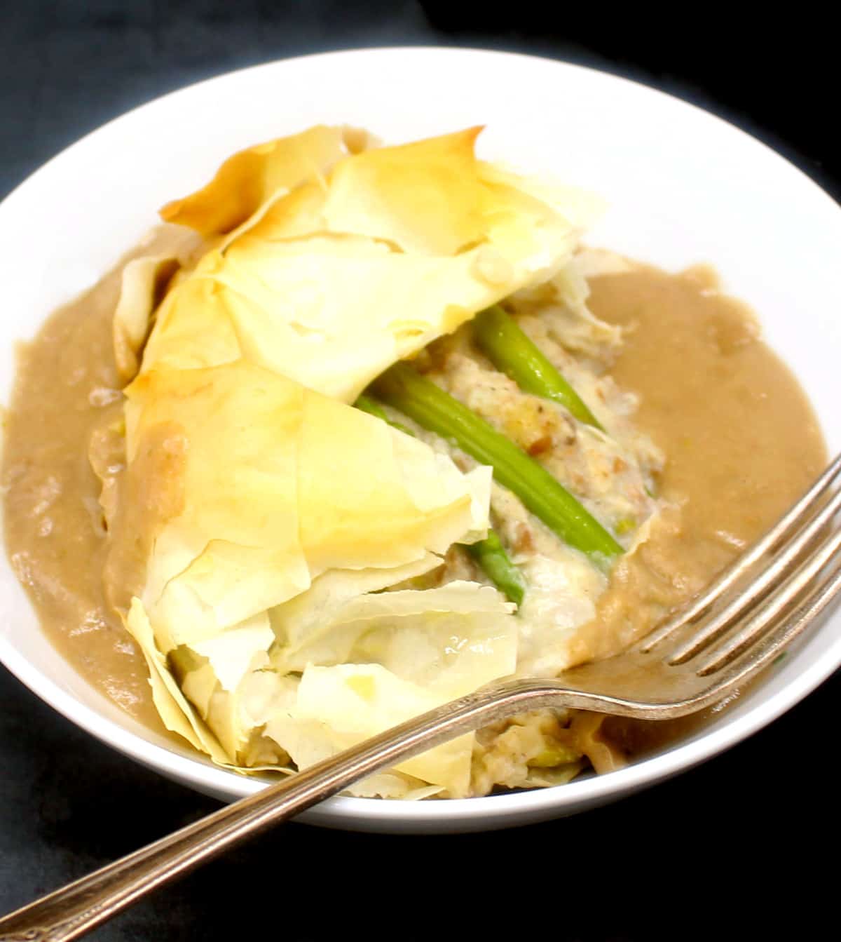A white bowl with a portion of vegan asparagus potato tart with vegan gravy and fork.