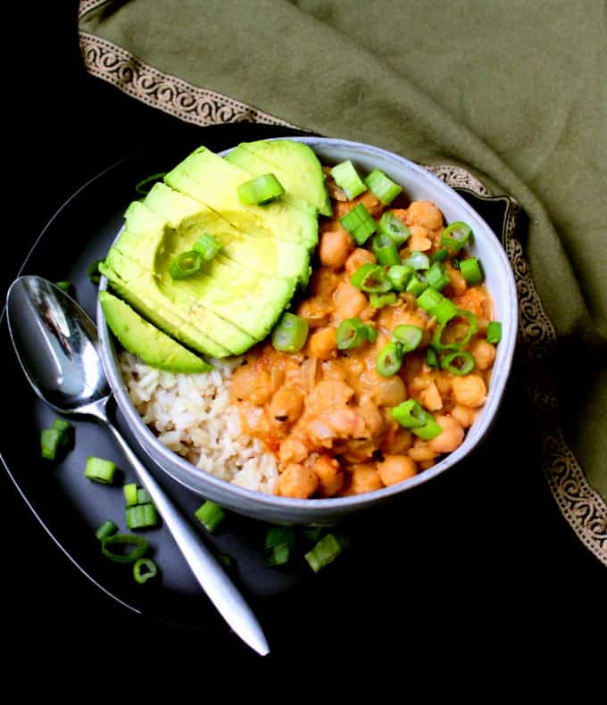 Vegan Thai curried chickpeas with vegan red curry paste in a glazed gray bowl with an avocad half, scallions and a silver spoon