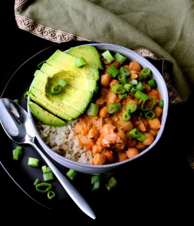 Vegan Thai curried chickpeas with vegan red curry paste in a glazed gray bowl with an avocad half, scallions and a silver spoon