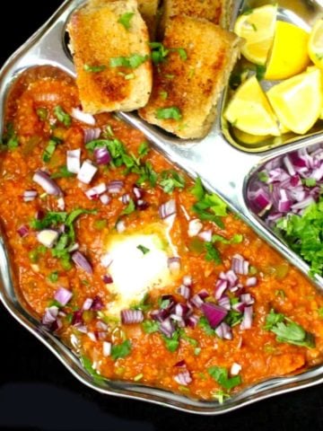 An overhead shot of a plate of pav bhaji with the mashed veggies, pav, lemons onions and cilantro
