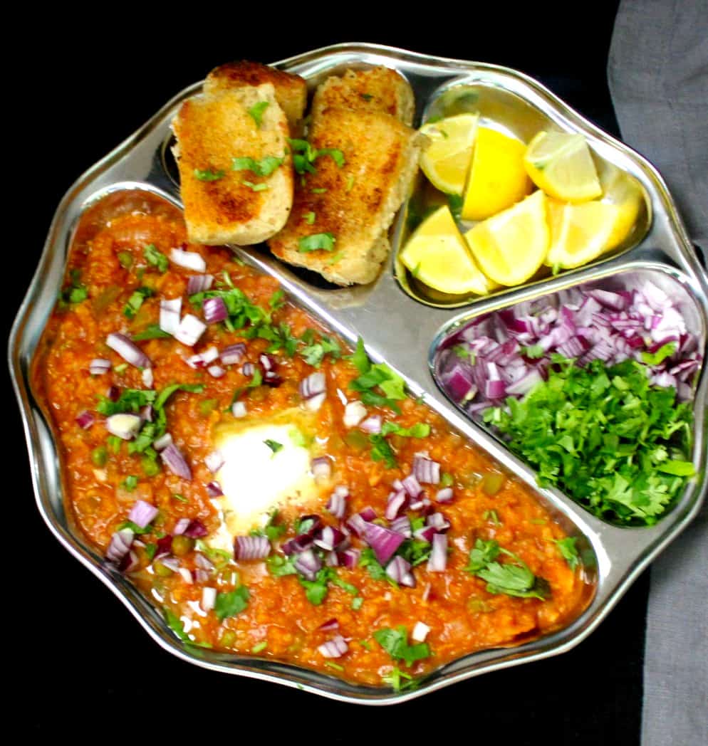 A front shot of pav bhaji in a steel plate with all the trimmings--onions, butter, lemon and cilantro on a black table.