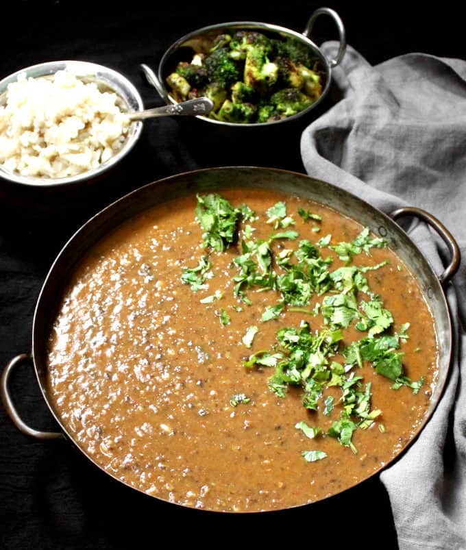 Een grote koperen pan met urad dal en koriander met bruine rijst en broccoli roerbak in de rug