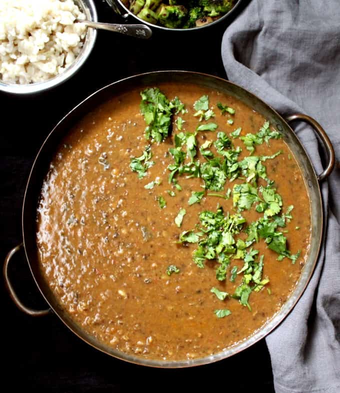 Spicy urad dal or black lentil dal, with creamy lentils and spiced with ginger, garlic and red pepper in a copper pan with cilantro, brown rice and a vegetable curry.