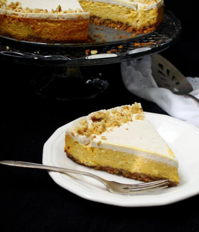 A slice of mango cheesecake with a graham-cracker-walnut crust on a white plate with a fork on the side and in the background is the full cake on a crystal cake stand with a silver serving spoon and a white lace napkin on the side.
