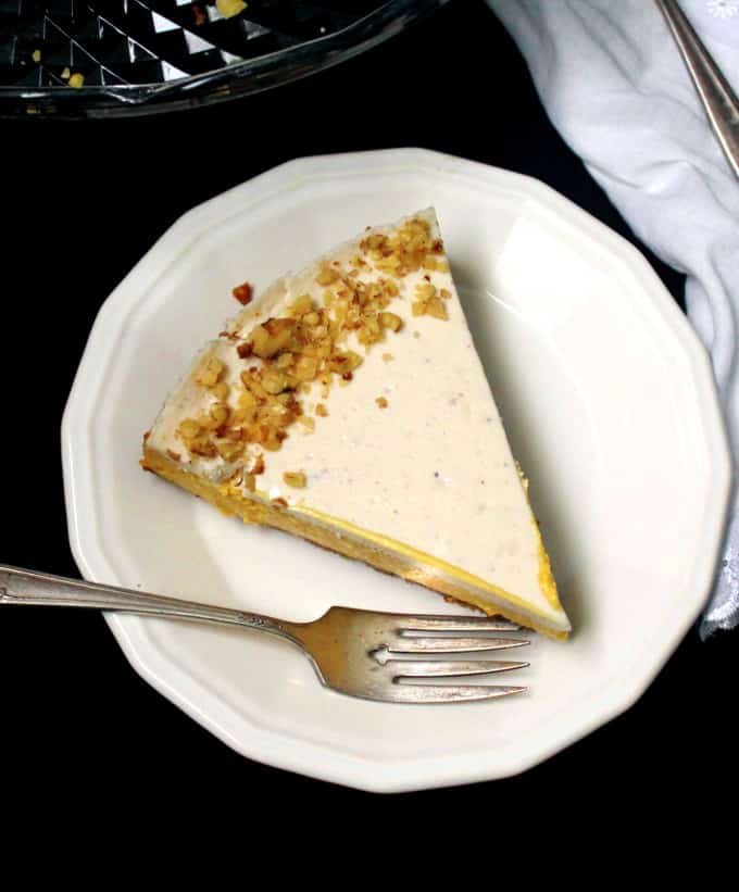 Top shot of a vegan mango cheesecake slice with walnuts scattered on top and sitting on a white plate with a decorative fork next to it, set against a black background.