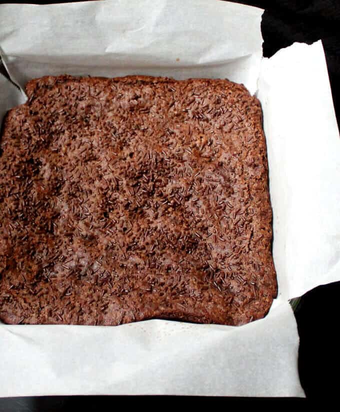 Closeup of baked vegan brownie with chocolate sprinkles in a square glass baking pan lined with parchment paper.