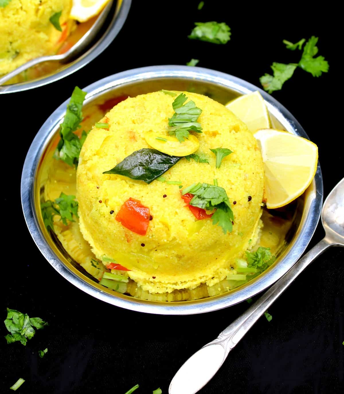 Upma with wedges of lemon, tomatoes, cilantro and curry leaves with a spoon next to it on a black background.