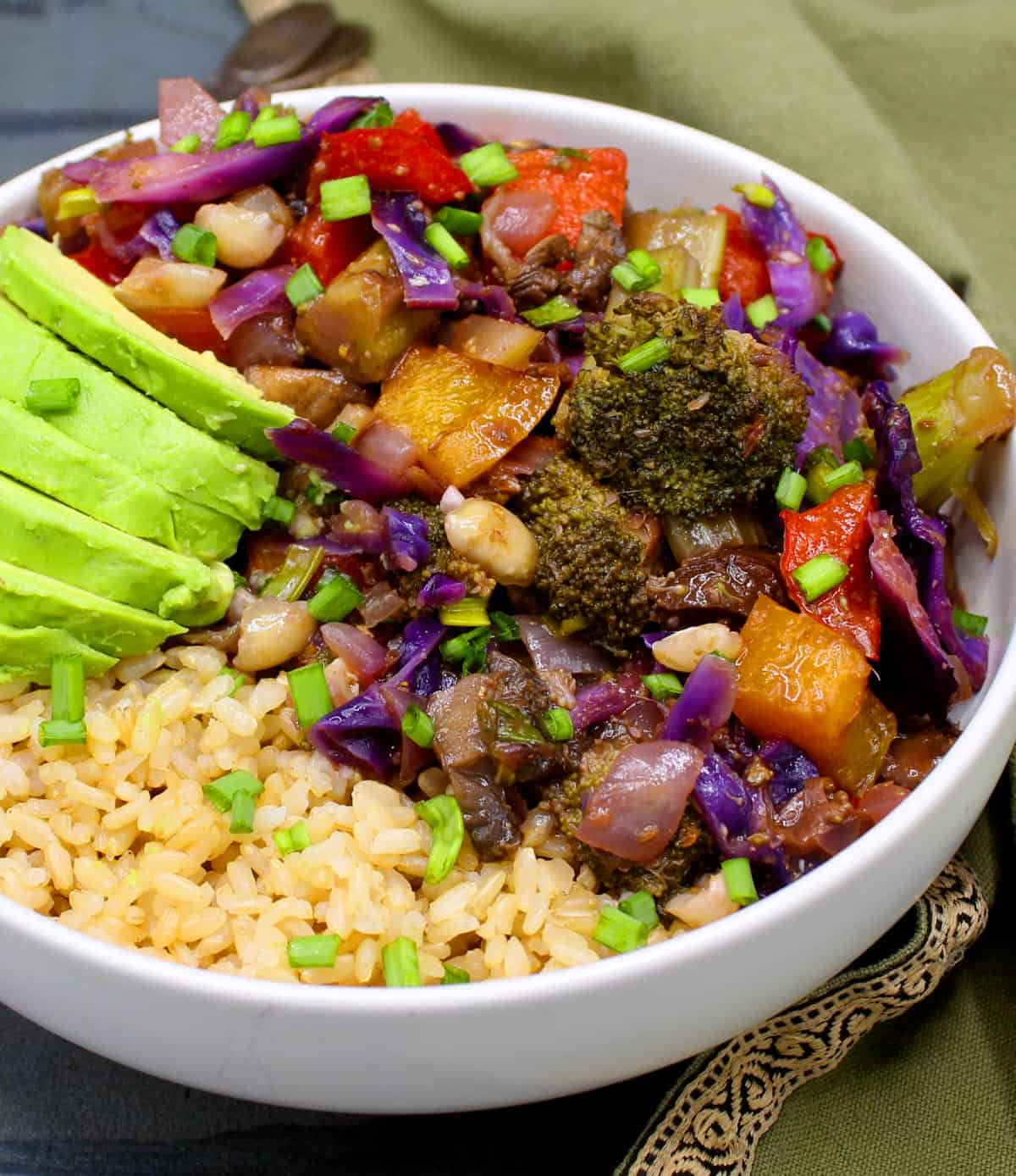 Kung pao vegetables served over rice in a bowl with avocado slices on the side.