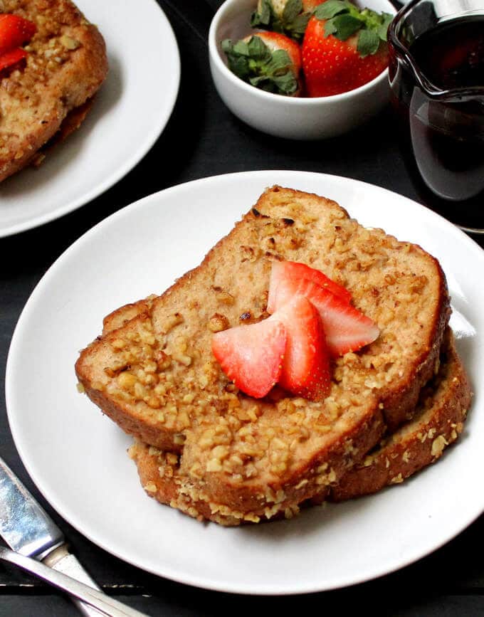 Two slices of vegan French toast with strawberries on a plate and maple syrup and more strawberries on the side.