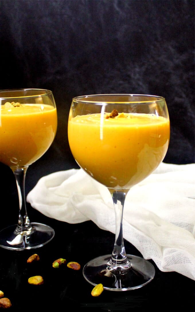 Two glasses of creamy vegan mango lassi garnished with pistachios with a cheesecloth nearby.