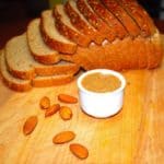 Homemade almond butter in small cup with almonds and a sliced bread in background.