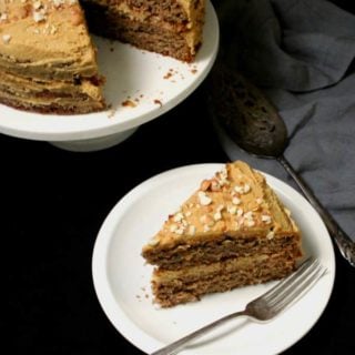 A top shot of a slice of vegan banana cake with peanut butter frosting on a white plate with a fork and the full cake on a cake stand in the back