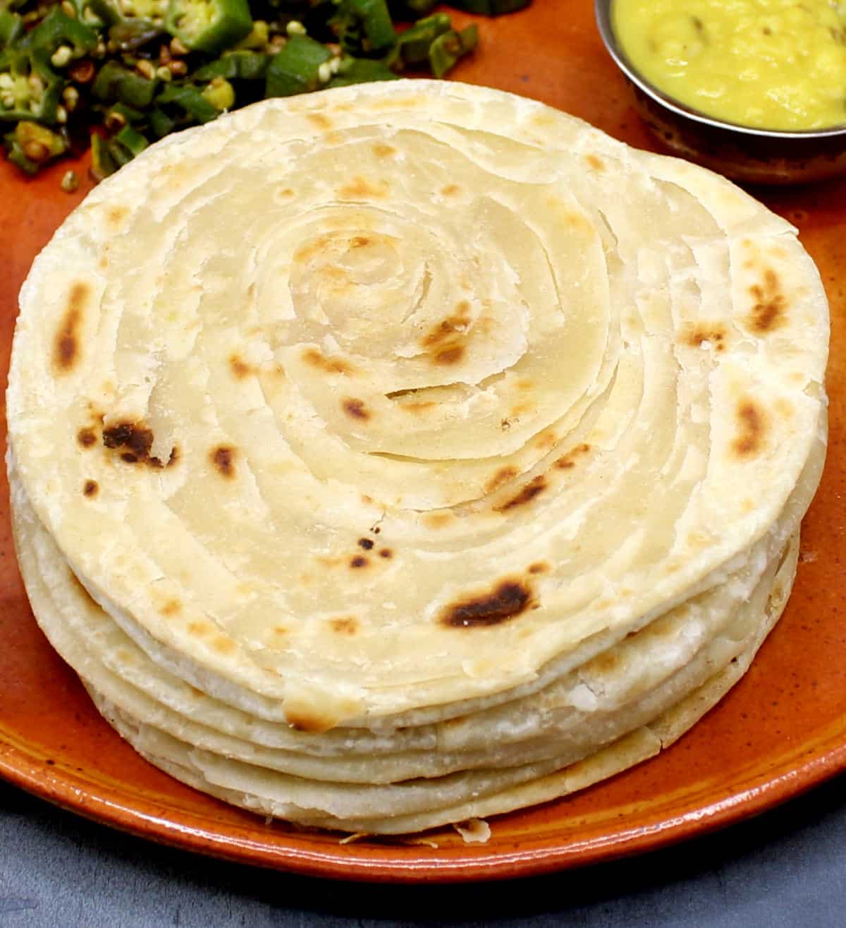 Parathas stacked on clay plate with dal and sabzi in background.