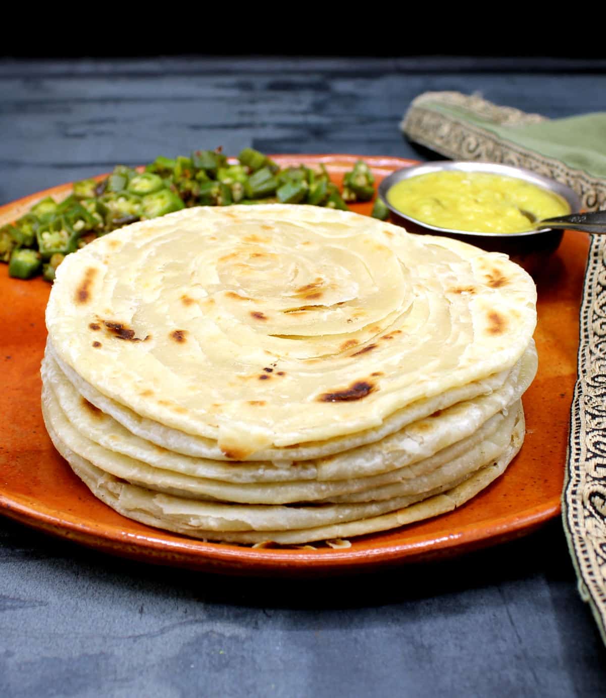 Flaky paratha stacked on clay plate with dal and sabzi.