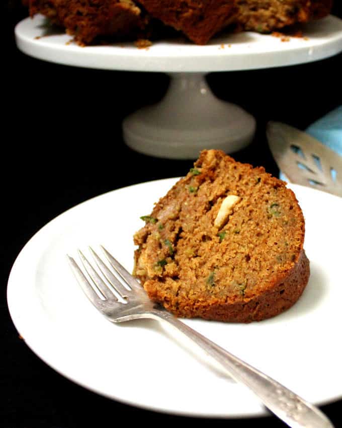 A slice of cake made with cucumbers, cashews, cardamom and whole wheat on a white plate with a silver fork