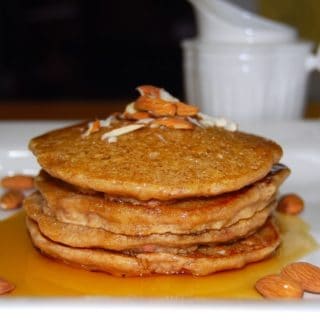 A stack of vegan almond pancakes topped with almonds and drizzled with maple syrup on white plate.