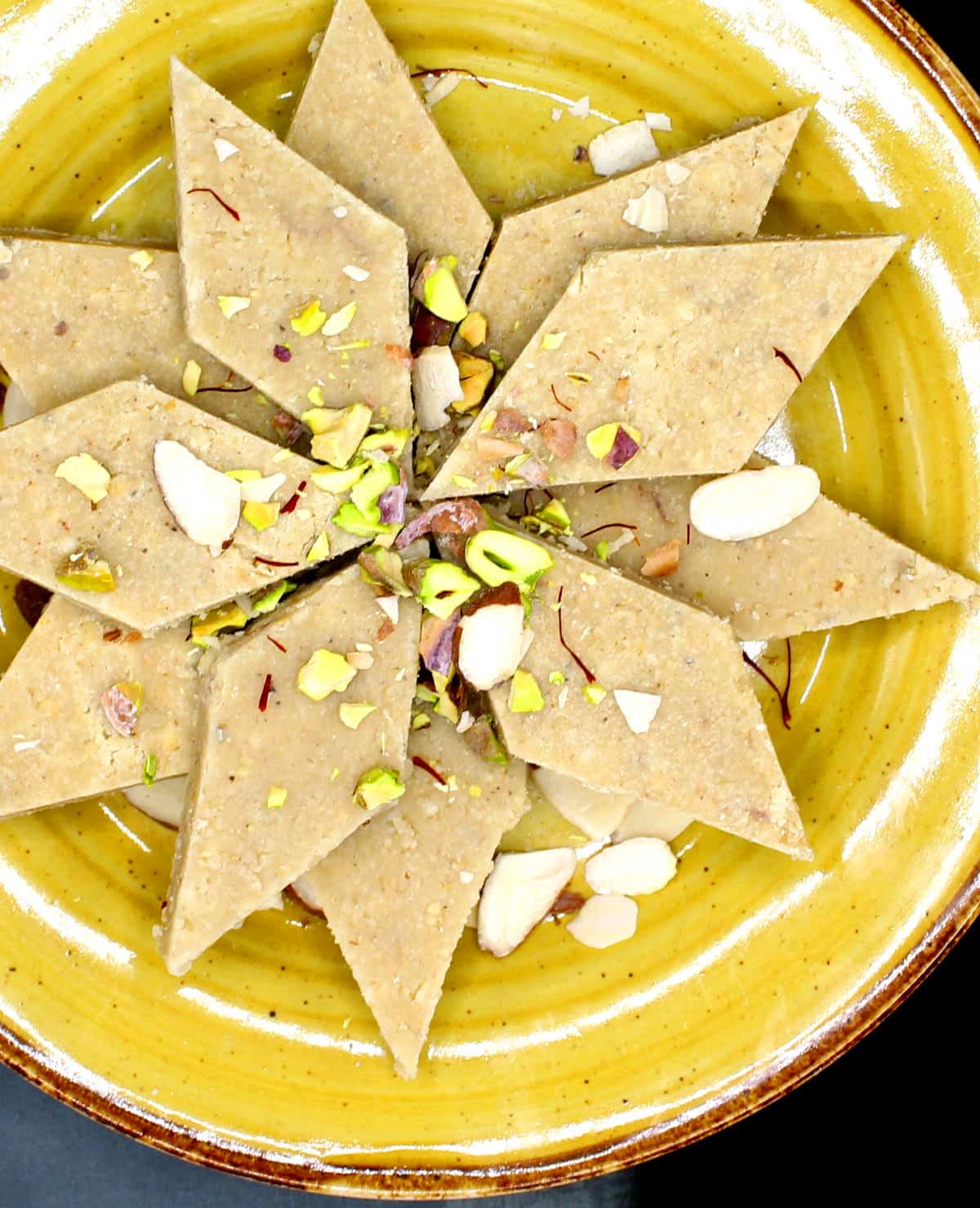 Vegan kaju katli arranged in yellow plate with nuts garnish.