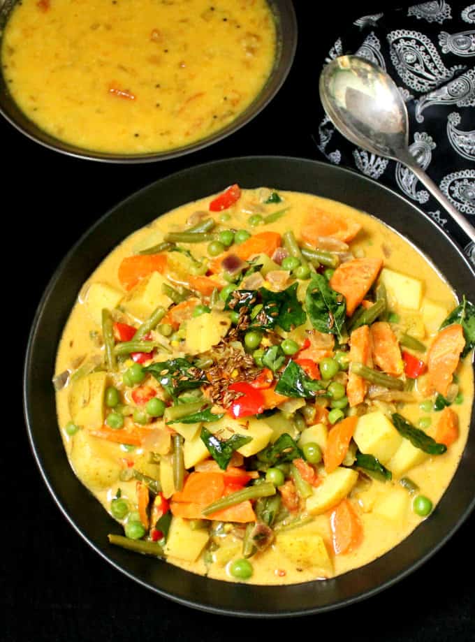 A bowl with vegetable korma, made with carrots, potatoes, green beans and tomatoes in a spicy but creamy coconut sauce. Next to the bowl is a paisley napkin and a bowl of dal and spoon.