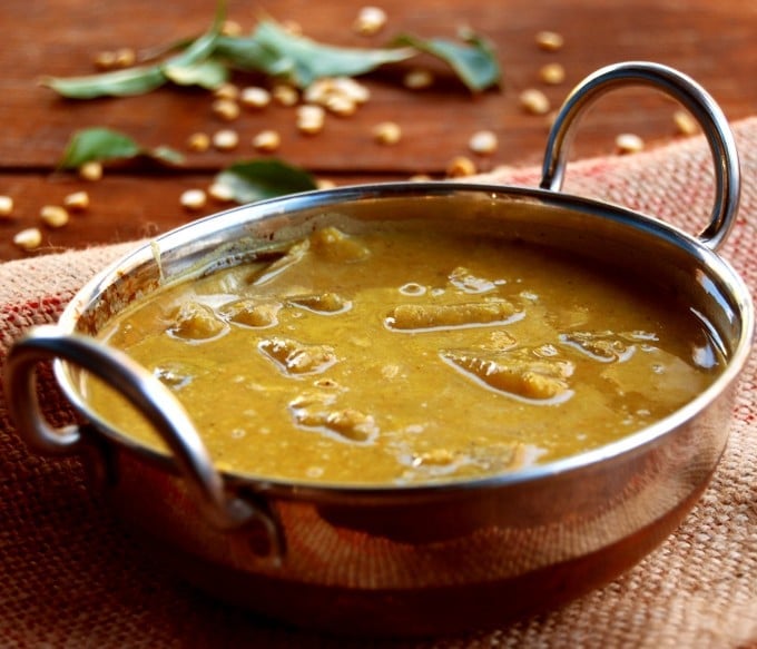 White pumpkin sambar in kadhai with spices, lentils and curry leaves in background.