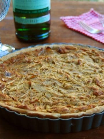 Vegan French onion tart in tart pan with wine and wine glasses in background.
