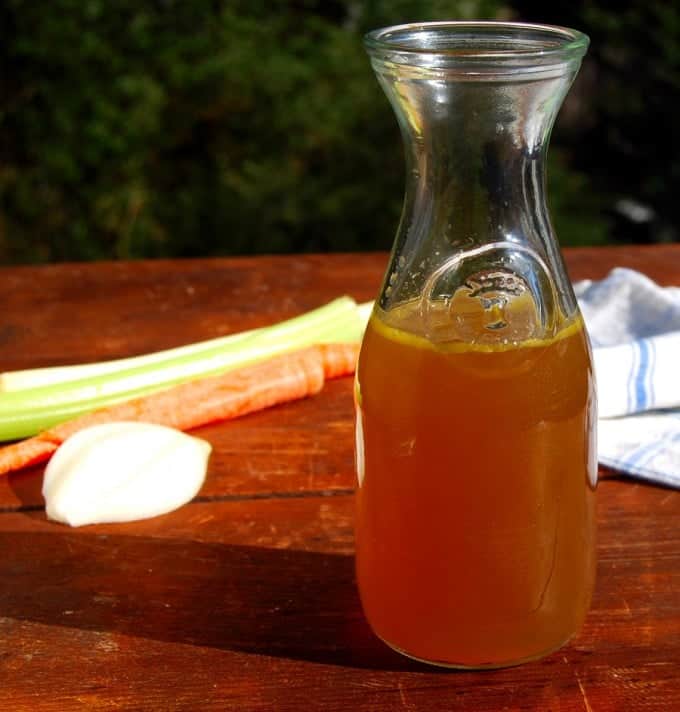 vegetable stock in glass container with vegetables like carrots and celery in background.