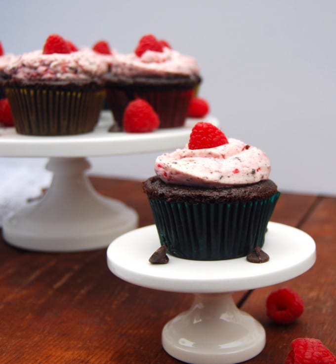 A vegan chocolate cupcake with raspberry frosting on cake stand with more cupcakes in background.