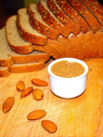 Homemade almond butter in small cup with almonds and a sliced bread in background.