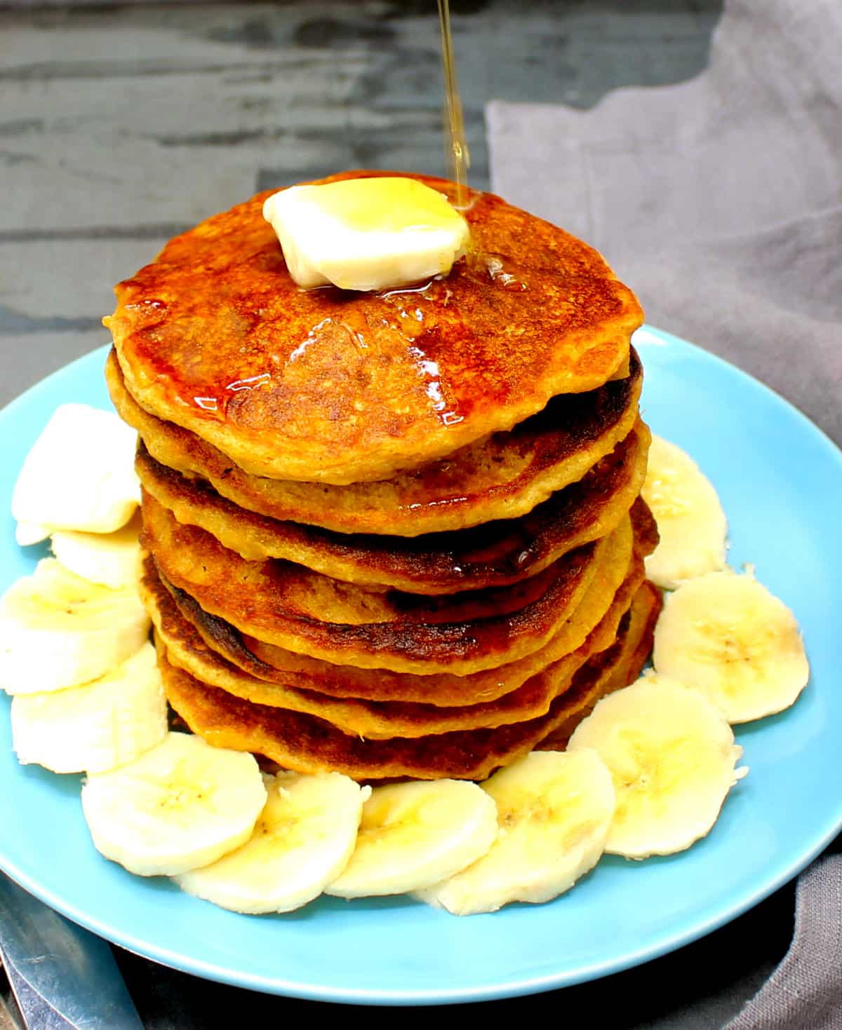 Vegan sweet potato pancakes in stack on blue plate surrounded by sliced bananas, with a pat of vegan butter on top and maple syrup poured over.