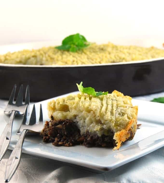 A slice of savory vegan lentil shepherd's pie with a base of lentils and mushrooms topped with a creamy layer of potatoes on a white plate with two forks