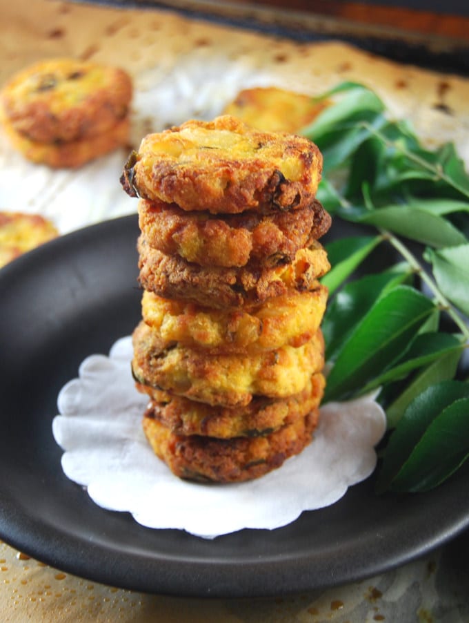 Baked, fat-free potato vadas (patties) stacked on a black plate with curry leaves.