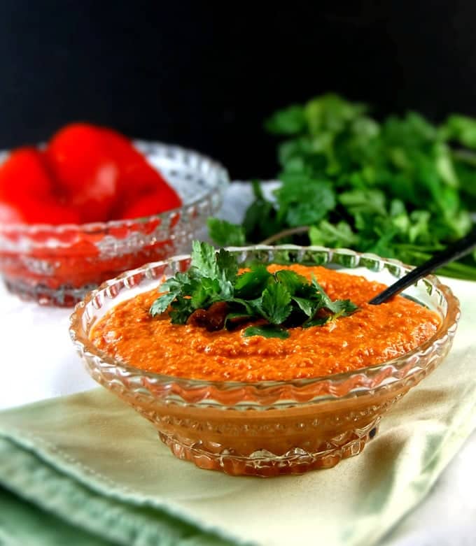 A crystal bowl with Roasted Red Pepper Chutney with cilantro garnish and a roasted red pepper in background.