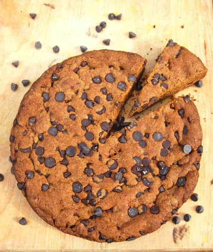 Overhead shot of a large vegan banana skillet cookie with a slice cut out on a wooden background with chocolate chips scattered around